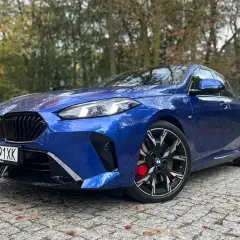Błękitne BMW 235i z czarnymi felgami i czerwonymi zaciskami hamulcowymi zaparkowane na bruku wśród jesiennych drzew.