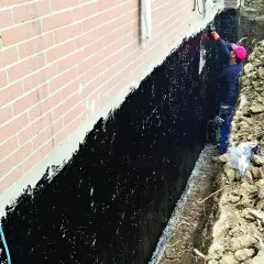 Pracownik nakłada czarną hydroizolację na ścianę fundamentową.