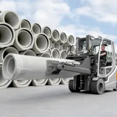 Wózek widłowy z operatorem transportuje dużą betonową rurę. W tle piętrzą się kolejne elementy tej jednostki ładunkowej.