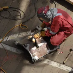 Spawacz w czerwonym kombinezonie pracuje nad metalowym elementem. Obok leży młotek i szlifierka, sugerując, jaki agregat do spawarki mógł być użyty.