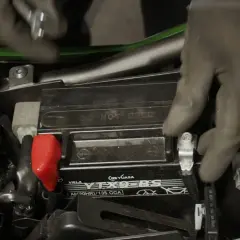 Manos enguantadas conectando la batería de una moto. Se ve el terminal positivo rojo y los cables.