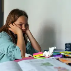 Mädchen sitzt gelangweilt am Schreibtisch, umgeben von Schulmaterialien. Endlich wieder Schule, aber die Motivation fehlt.