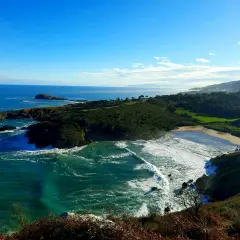 Vista panorámica de la playa de Sonabia, con olas rompiendo en la costa rocosa y un mar azul intenso bajo un cielo despejado.