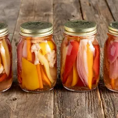 Quattro barattoli di giardiniera fatta in casa, con verdure colorate come peperoni, carote e cavolfiore, su un tavolo di legno rustico.