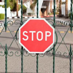 Czerwony znak STOP na zielonym, ozdobnym płocie. Symbolizuje problemy ze służebnością drogi, blokując dostęp.