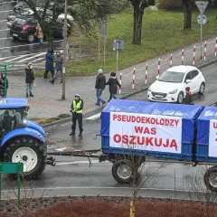 Niebieski traktor ciągnie przyczepę z banerami "Pseudoekolodzy was oszukują" i "Unijna polityka zniszczy rolnika".
