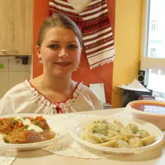 Uśmiechnięta kobieta w tradycyjnej koszuli prezentuje dania: gołąbki, pierogi i zupę. Pyszny, domowy obiad jak u Tarasa Bulby.