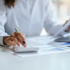 Osoba liczy na kalkulatorze, przegląda dokumenty i laptopa. Przygotowuje się do złożenia karty podatkowej.