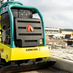 Robotnik na budowie, obok niego stoi żółta zagęszczarka Ammann. Wygląda na to, że zagęszczarka nie jedzie do przodu, bo operator stoi obok.
