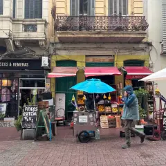 Un hombre con capucha camina por una calle adoquinada en Montevideo, pasando por tiendas de discos y un puesto de frutas.