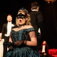 Kobieta w maskę i tiarze śpiewa na scenie. W tle mężczyźni w smokingach. Spektakle w Teatrze Gdynia.