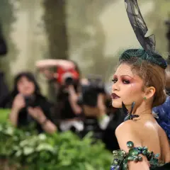 Una modelo con un elaborado atuendo de **Met Gala theme**, con velo y maquillaje dramático, posa rodeada de fotógrafos y vegetación.