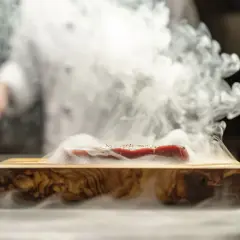 Un morceau de viande rouge est posé sur une planche en bois, entouré d'une fumée dense. Cela illustre comment fonctionne un fumoir pour infuser des arômes.