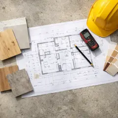 Bauplan, Materialmuster und Farbpalette für die Sanierung eines Hauses. Gelber Helm und Messgerät deuten auf die Kosten hin.