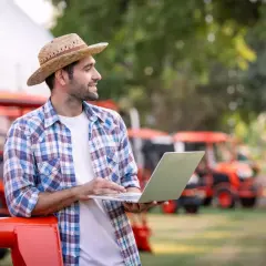 Rolnik w kapeluszu słomkowym z laptopem na tle czerwonych traktorów. Zastanawia się, jak przejść na krus, planując przyszłość gospodarstwa.