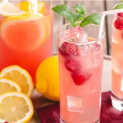 Två glas med rosa lemonad, hallon, is och mynta. Citroner och en karaff i bakgrunden ger härliga sommar vibes.