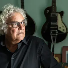 Massimo Bubola, con i suoi capelli ricci e bianchi, guarda pensieroso tra le chitarre appese al muro, testimoni silenziose delle sue canzoni.