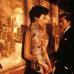 In the mood for love spiegazione: una donna in cheongsam fiorito e un uomo in abito scuro si guardano con intensità, sullo sfondo di un muro con scritte cinesi.