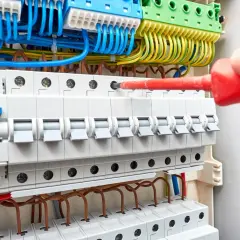 Ręka z czerwonym śrubokrętem przykręca przewody w skrzynce elektrycznej. Trwa wymiana instalacji elektrycznej.