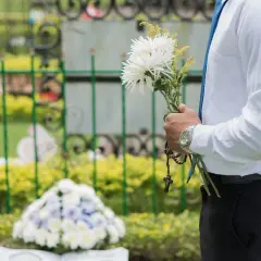 Mężczyzna w białej koszuli i niebieskim krawacie trzyma bukiet białych kwiatów i różaniec. W tle widać nagrobki i zielone krzewy. Msza pogrzebowa z urną trwa.