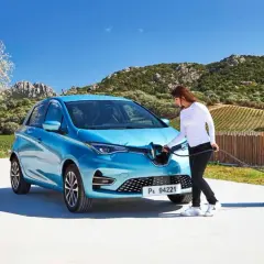 Une femme branche sa Renault Zoé bleue à une borne de recharge. Le temps de recharge zoe 22kw est rapide, parfait pour profiter du paysage.