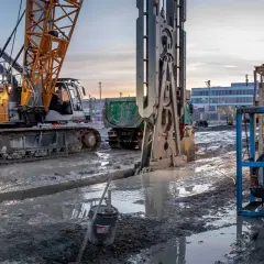 Baustelle bei Dämmerung: Kran und Bohranlage im Einsatz für die Schlitzwand Herstellung. Schlammige Gräben und schwere Maschinen prägen das Bild.