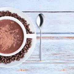 Tazza di cioccolata calda con cacao amaro in polvere e chicchi di cioccolato, su un piatto bianco, accanto a un cucchiaino. Le proprietà del cacao amaro sono note per il benessere.