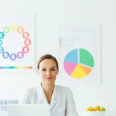 Dietetyczka w białym kitlu przy biurku, na ścianie wykresy z witaminami i makroelementami.