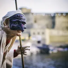 Pulcinella con maschera viola, pronto a litigare in napoletano, con Castel dell'Ovo sullo sfondo.