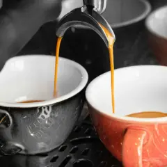 Dwa kubki, jeden szary, drugi pomarańczowy, napełniają się gorącym, aromatycznym espresso doppio.