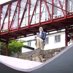 Estatua de un hombre sentado frente a la fábrica de Sargadelos, bajo un puente rojo y acristalado.