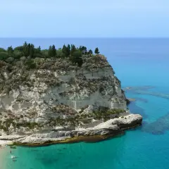 Scogliera bianca con un santuario in cima, bagnata da un mare cristallino in Calabria. Spiaggia affollata e barca all'orizzonte.