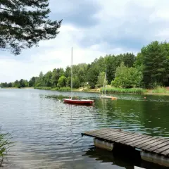Spokojne jezioro, dwa żaglówki na wodzie, drewniany pomost i zalesione **chechło** w tle.