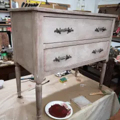 Un mobile antico in fase di restauro, con cassetti e pomelli decorati. Attrezzi e materiali da lavoro circondano il pezzo.