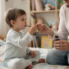 Mama podaje dziecku syrop z dekstrometorfanem. Maluch siedzi na łóżku w piżamce, a mama trzyma buteleczkę i łyżeczkę.