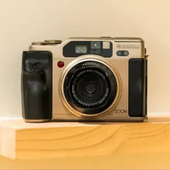 A gold Fujifilm 645 camera with a black grip sits on a wooden shelf.