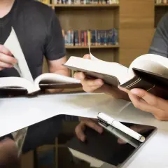 Estudiantes consultan libros y tablet en biblioteca, preparándose para las competencias específicas LOMLOE Andalucía.