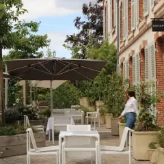 Terrasse verdoyante du 25bis by Leclerc Briant, avec tables, chaises et parasols. Une femme se dirige vers la réception.