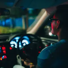 Un hombre con gafas conduce de noche, con el tablero iluminado en azul y rojo.
