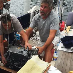 Due uomini lavorano alla manutenzione del motore di una barca a vela, sollevandolo con una catena.