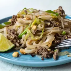 Pyszny pad thai przepis na talerzu: makaron ryżowy z mielonym mięsem, cebulą, orzeszkami ziemnymi i limonką.