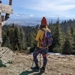 Widok na Tatry z drewnianego słupa z napisem "Koliba na Łapsowej Polanie". Turystka w kolorowych legginsach podziwia panoramę.