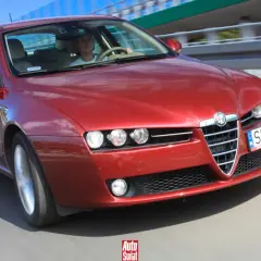 Czerwone Alfa Romeo 159 1.9 JTD pędzi po drodze. Kierowca w białej koszulce pewnie trzyma kierownicę.