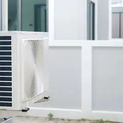 Unité extérieure d'une pompe à chaleur appartement, fixée sur un mur blanc. L'appareil est blanc avec des grilles bleues et un ventilateur visible.