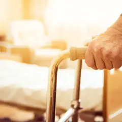 Dłoń osoby starszej chwyta chodzik. W tle łóżko szpitalne, sugerujące potrzebę wsparcia i dodatek mieszkaniowy dla osób niepełnosprawnych.