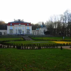 Pałac w Brzesku Okocimiu z ogrodem i alejkami. Słońce oświetla trawnik.