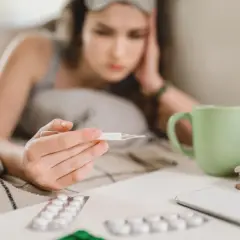 Chora dziewczyna z termometrem w ręku, leżąca w łóżku. Obok leki i kubek herbaty. L4 online 49 zł.