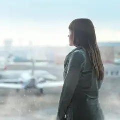 Dziewczyna w szarym płaszczu patrzy przez okno na lotnisku, gdzie w oddali widać samolot Boeing 737-8.