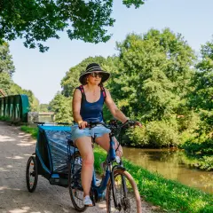 Mujer pedalea por las vías verdes de Francia con un remolque para niños, disfrutando del paisaje junto al canal.