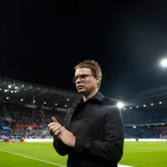 Właściciel Wisły Kraków, w czarnym płaszczu, stoi na murawie stadionu, patrząc w bok.